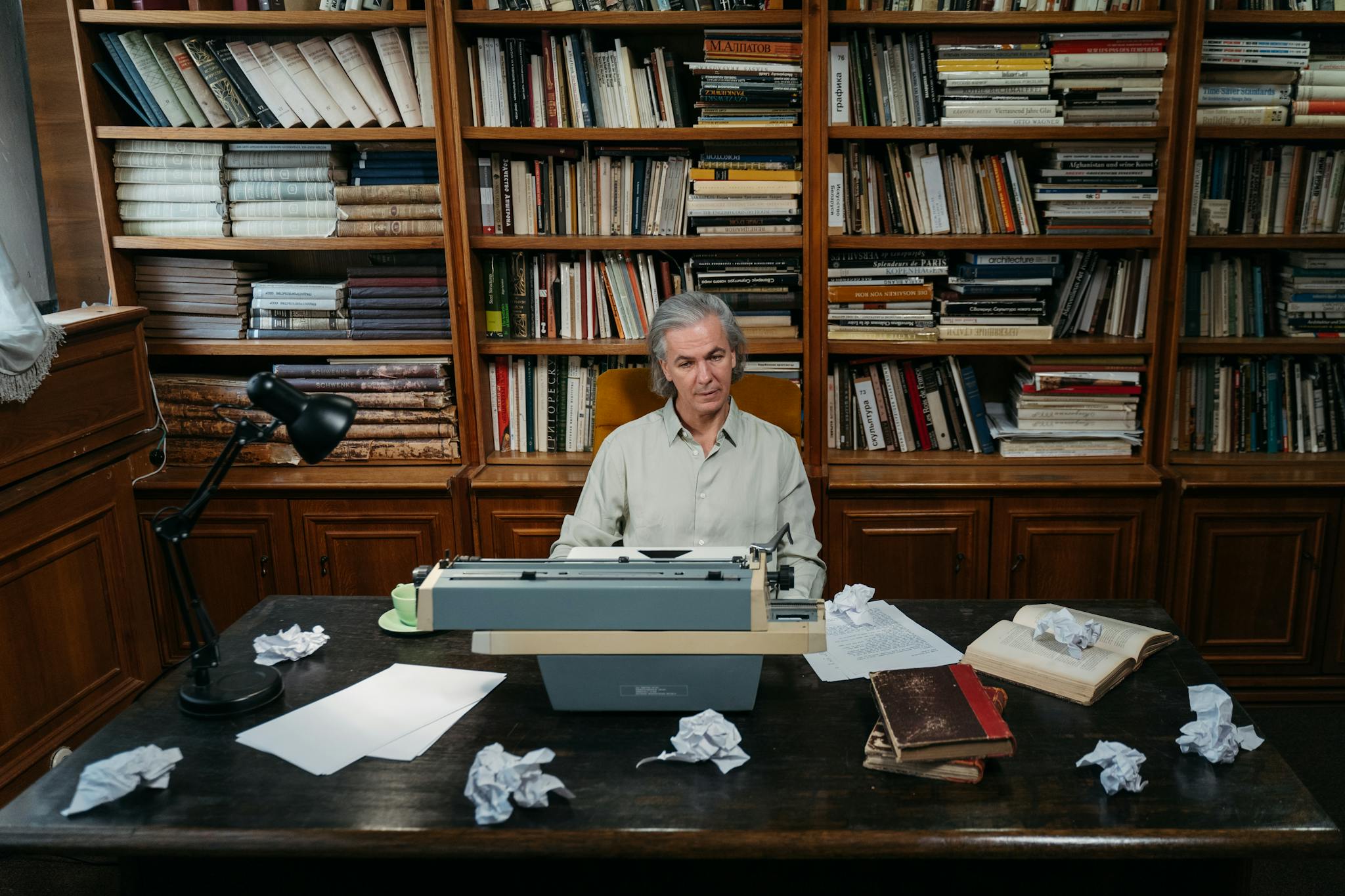 Senior man at a typewriter in a library, surrounded by books and paper.